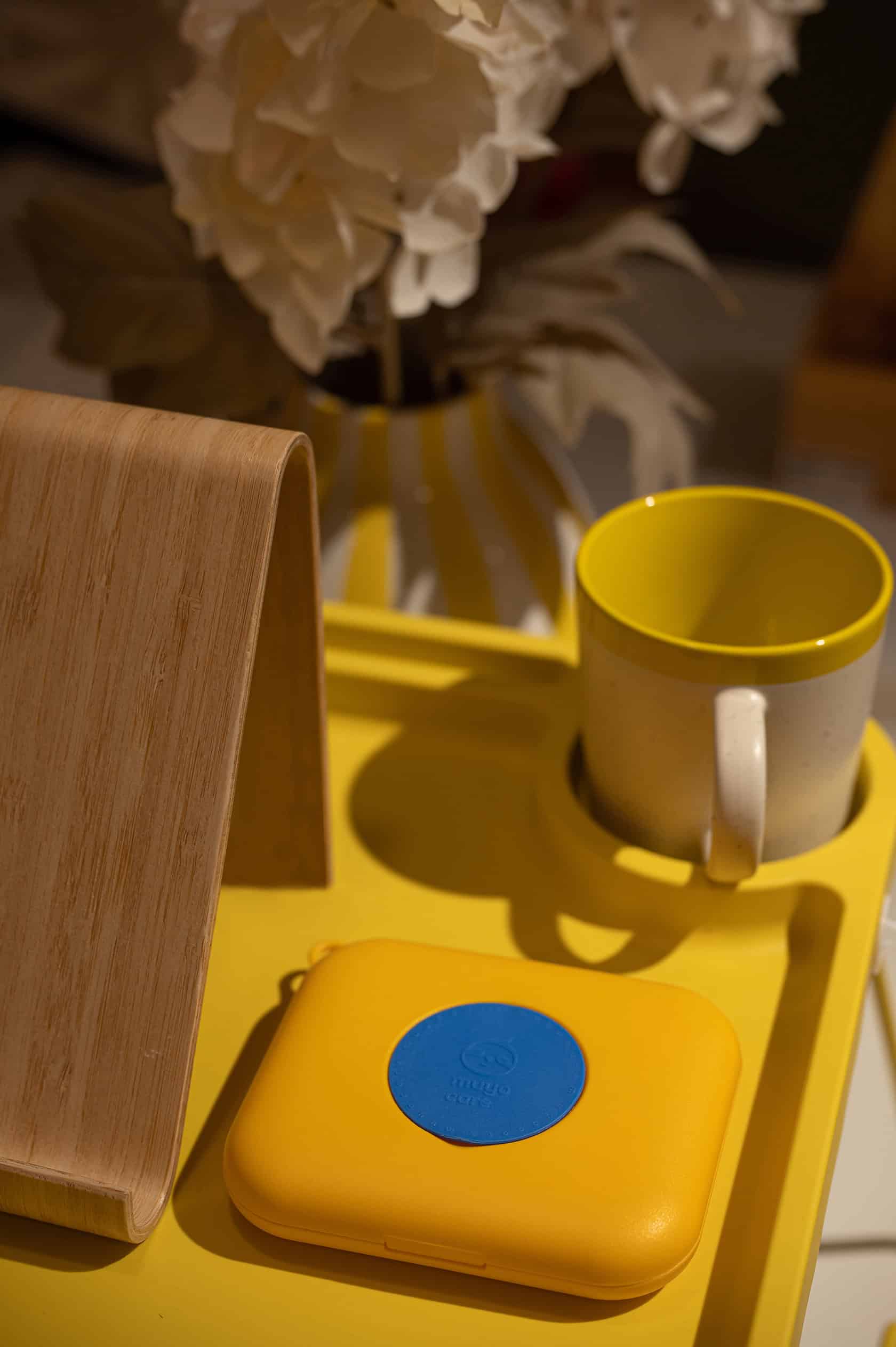 Bright yellow tray with MuyoCare device, ceramic mug, wooden stand, and hydrangea arrangement.